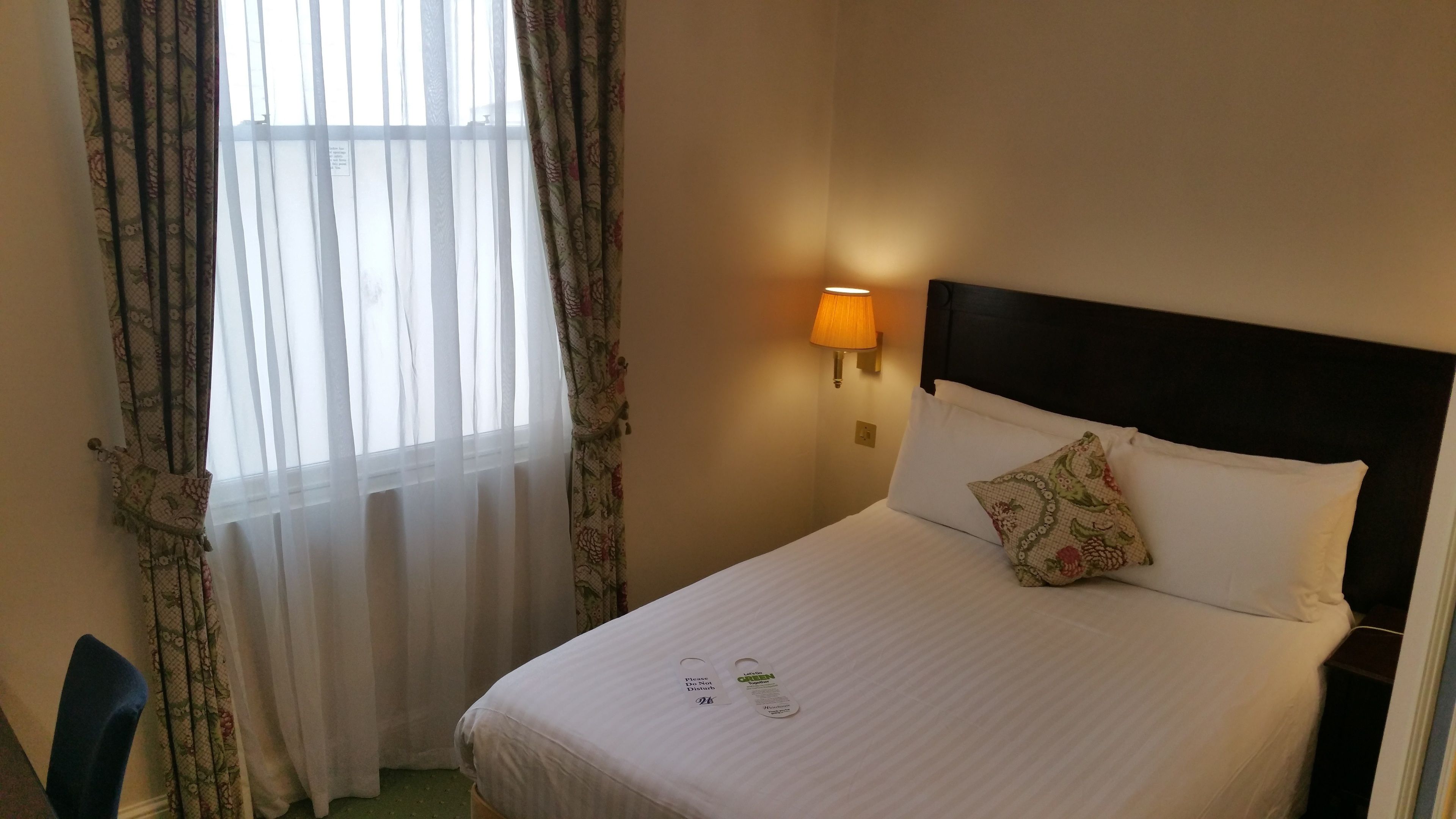 Ett hotellrum med en säng, ett nattduksbord, en lampa och ett fönster med gardiner.