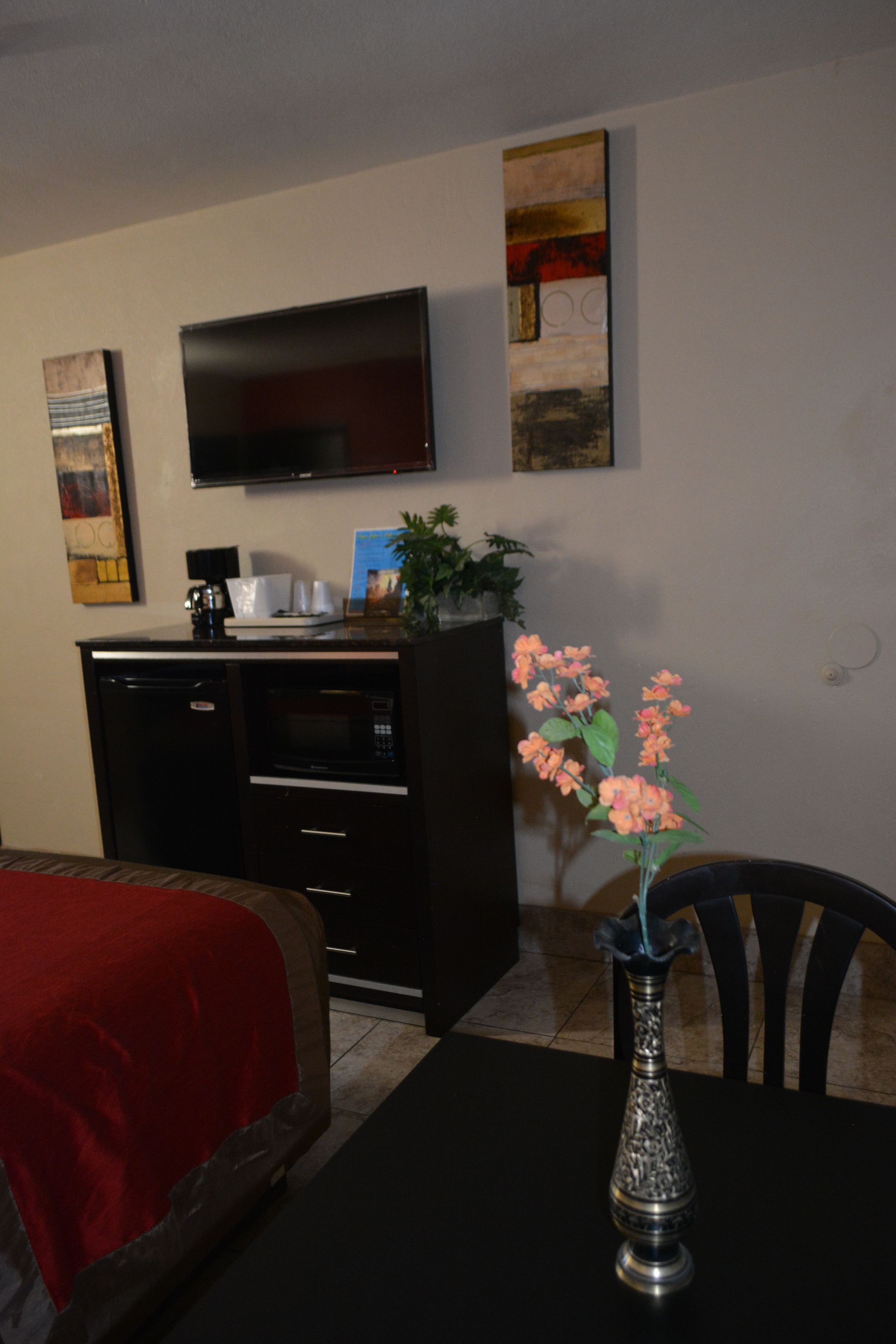 A hotel room with a dining table, a red sofa, a black cabinet with a flat-screen TV, a decorative vase with flowers, and a wall-mounted artwork.
