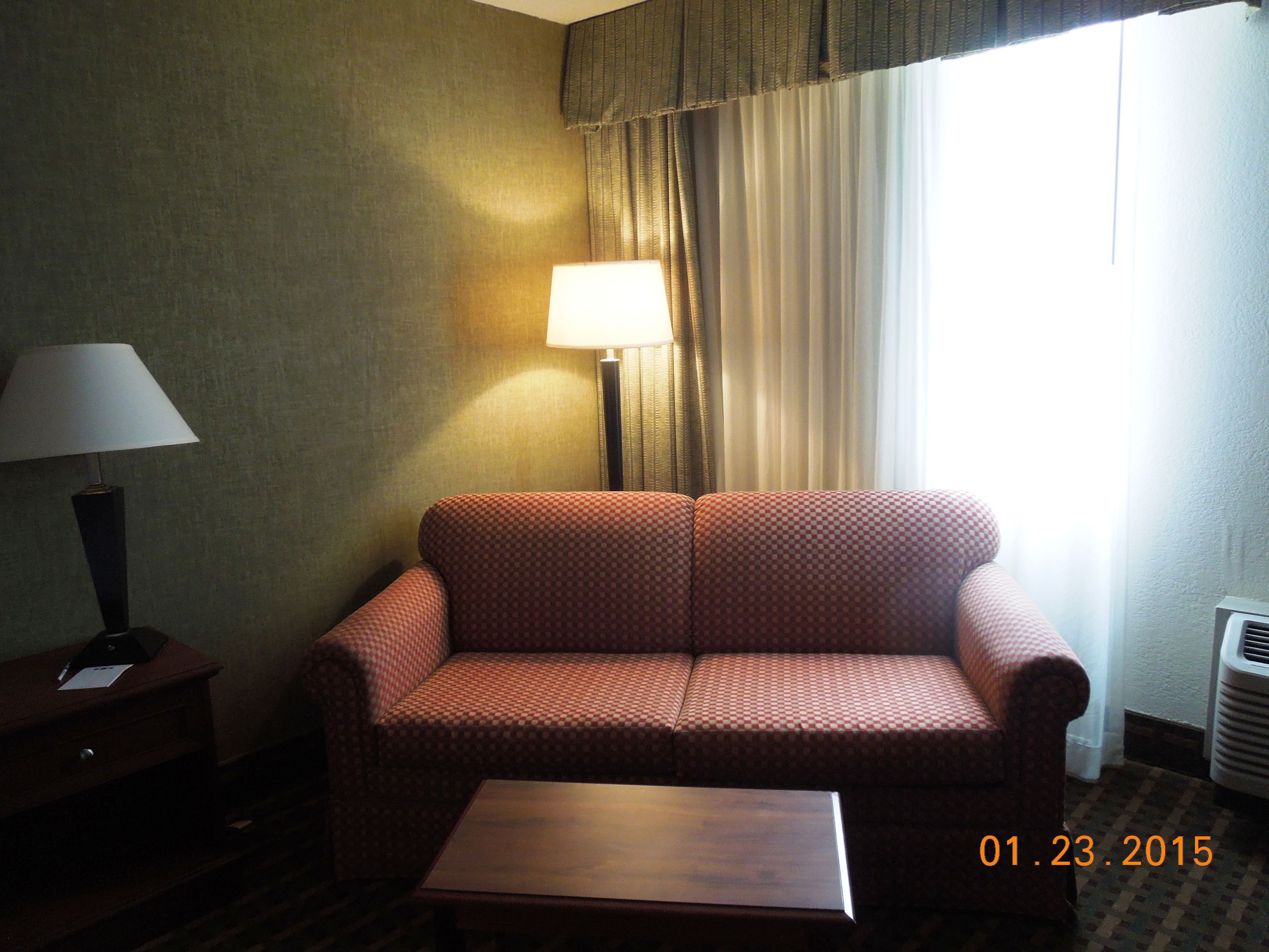 Habitación de hotel con un sofá a cuadros, una mesita, una lámpara y una ventana con cortinas.