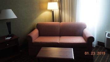 Habitación de hotel con un sofá a cuadros, una mesita, una lámpara y una ventana con cortinas.