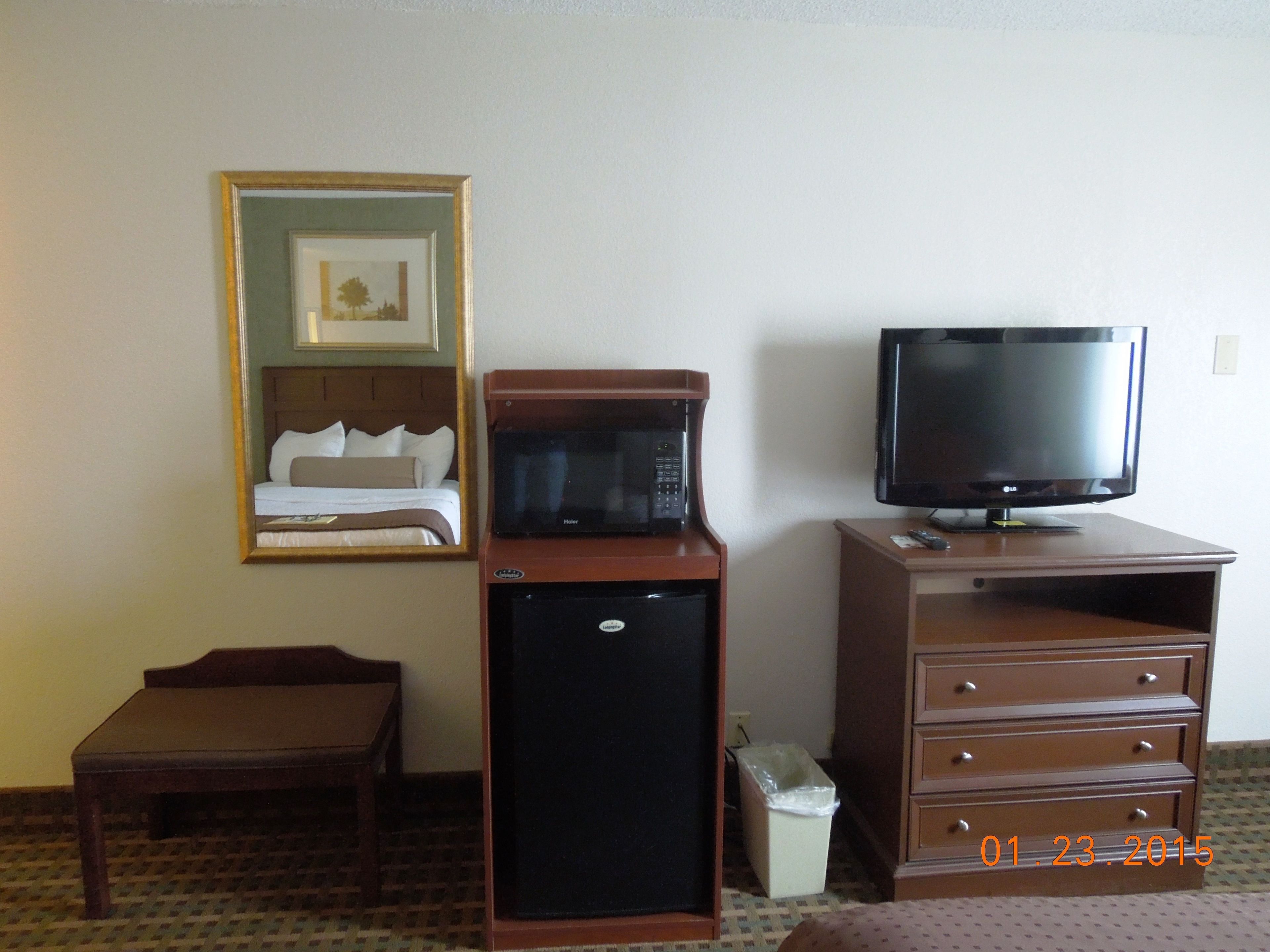 A hotel room with a bed, a microwave, a television, a small refrigerator, a trash bin, and a wooden bench.
