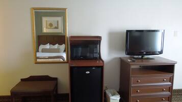 A hotel room with a bed, a microwave, a television, a small refrigerator, a trash bin, and a wooden bench.