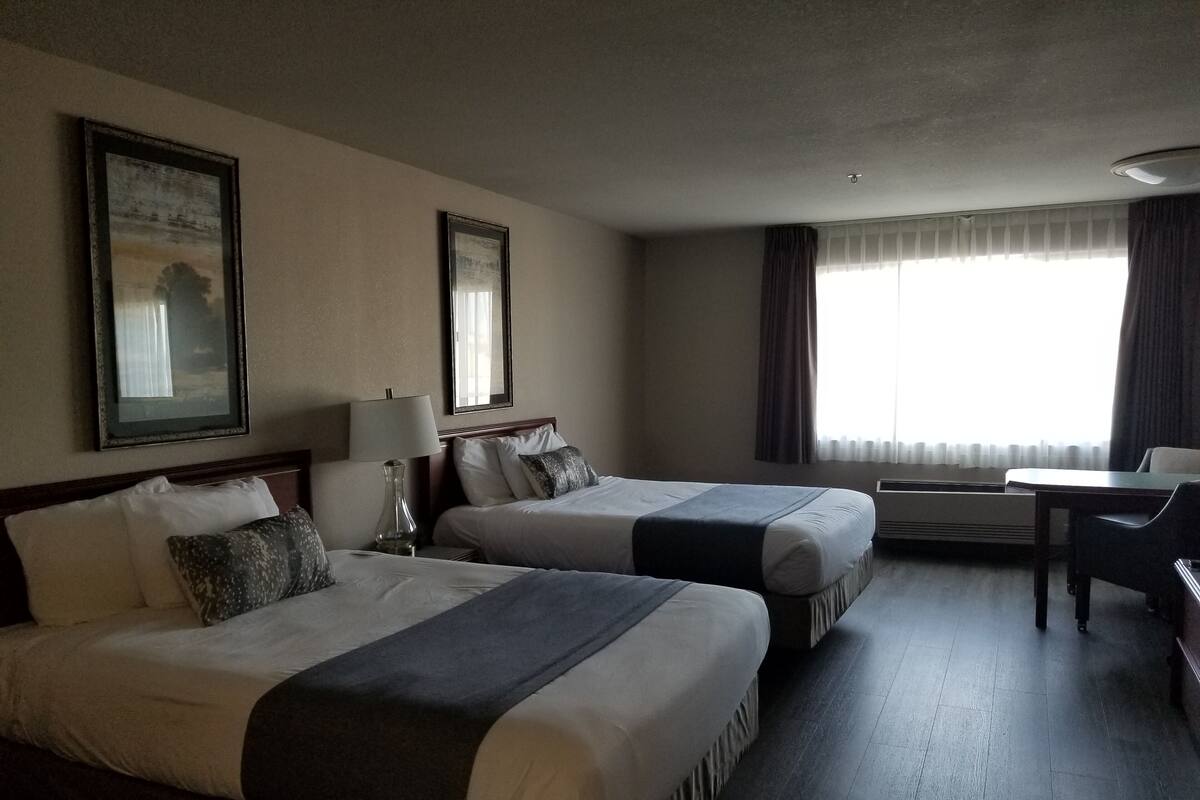 A hotel room with two beds, a desk, a chair, a lamp, and a window with curtains.