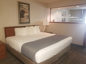 A hotel room with a bed, a nightstand, a lamp, a framed picture on the wall, and a window with a view of a building.