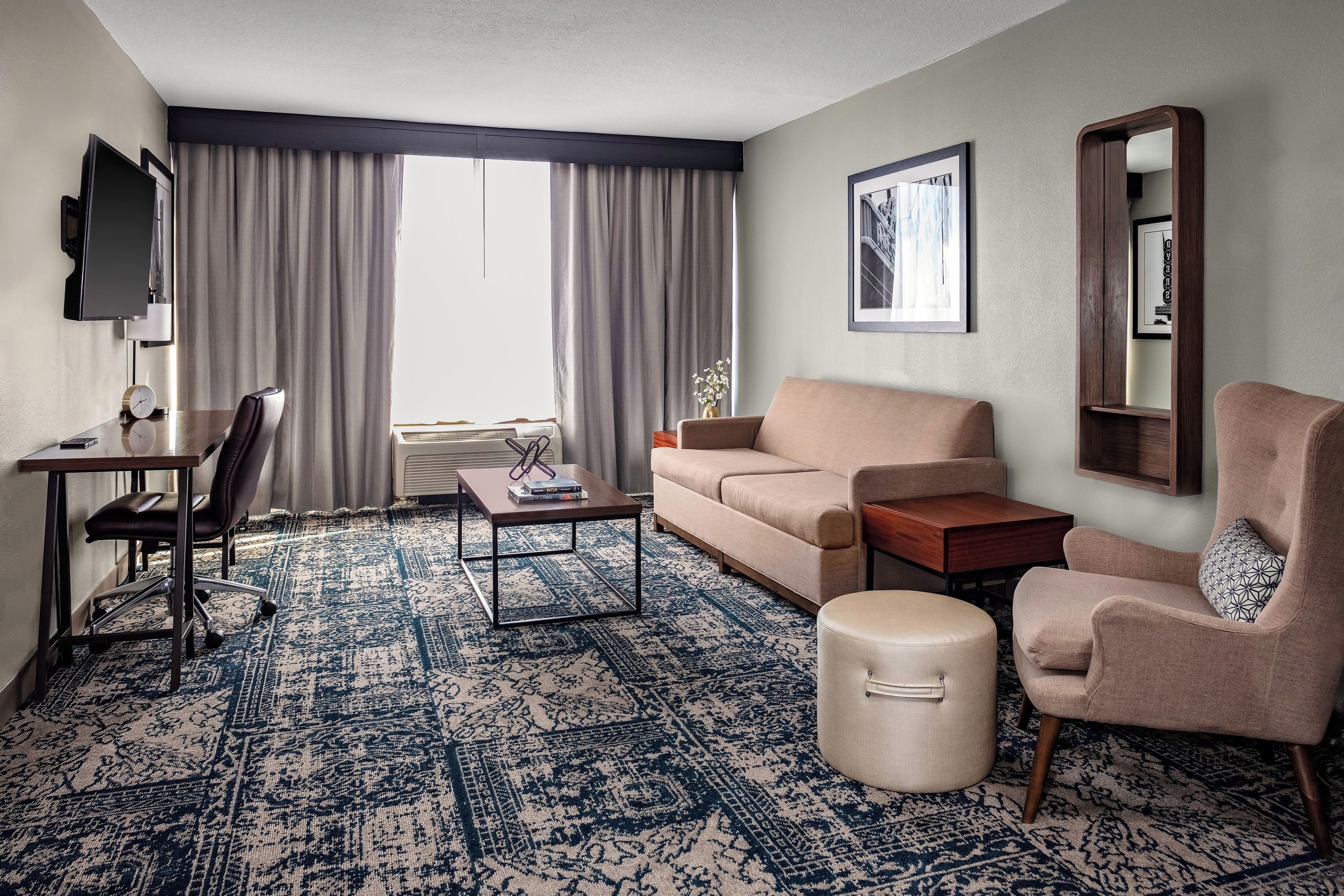 Quarto de hotel com sofá, poltrona, mesa de centro, televisão e uma grande janela com cortinas.