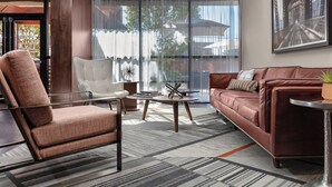 A modern living room with a brown leather sofa, a white armchair, and a brown armchair. There is a large window with sheer curtains, a painting on the wall, and a small table with a book and a decorative item. - Four Points by Sheraton Memphis East (Memphis)