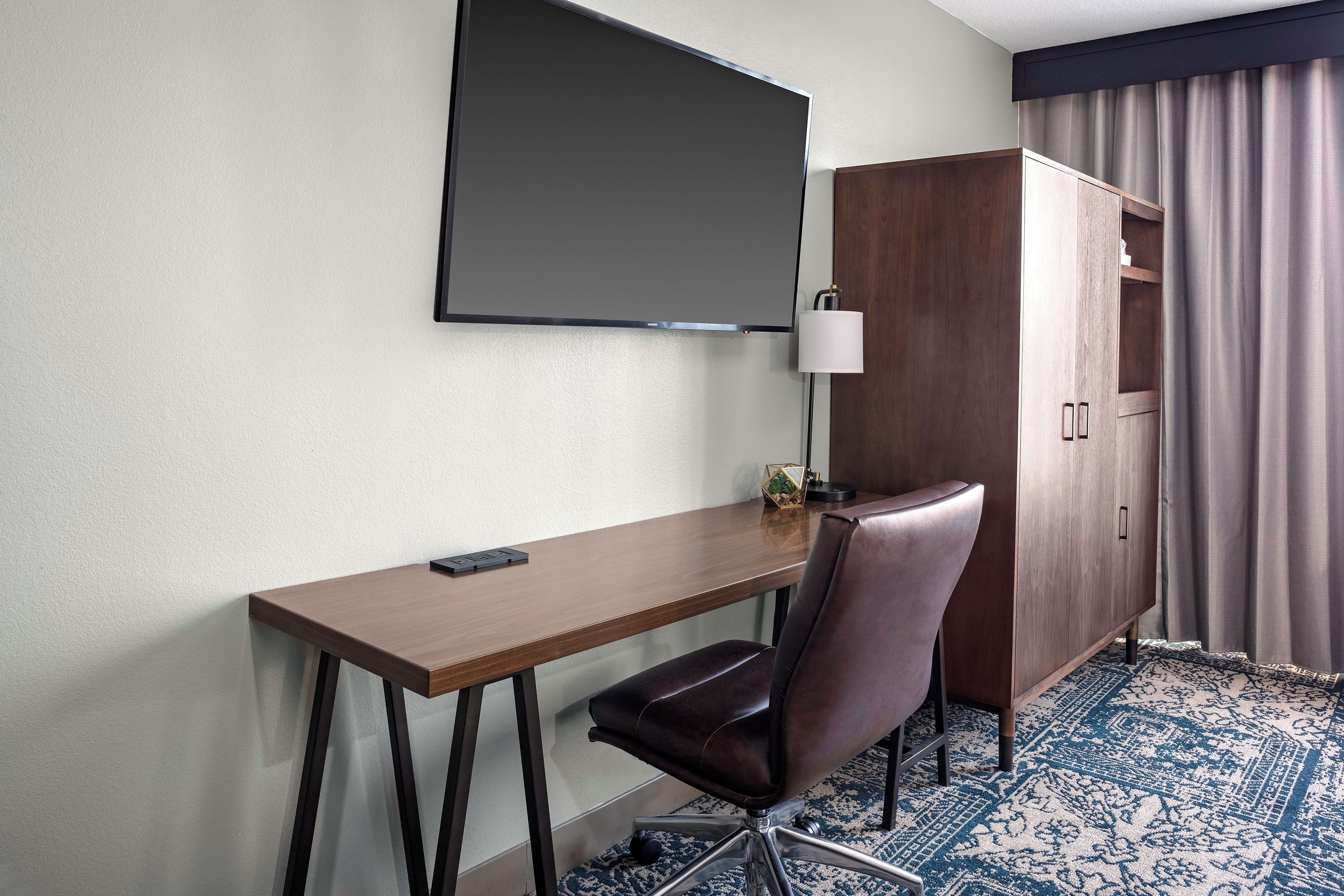 Habitación de hotel con escritorio, silla, televisor, armario y alfombra estampada.