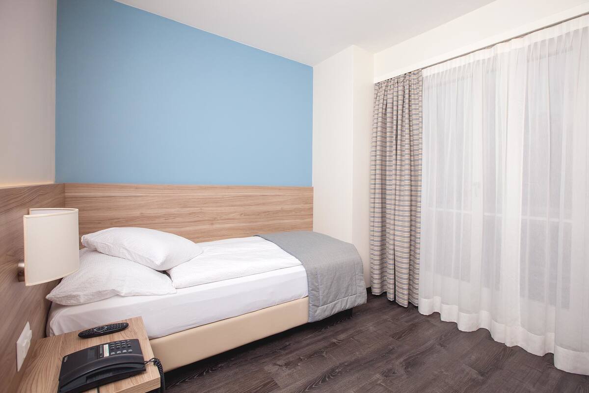 A hotel room with a bed, a bedside table with a lamp, a phone, and a window with sheer curtains.