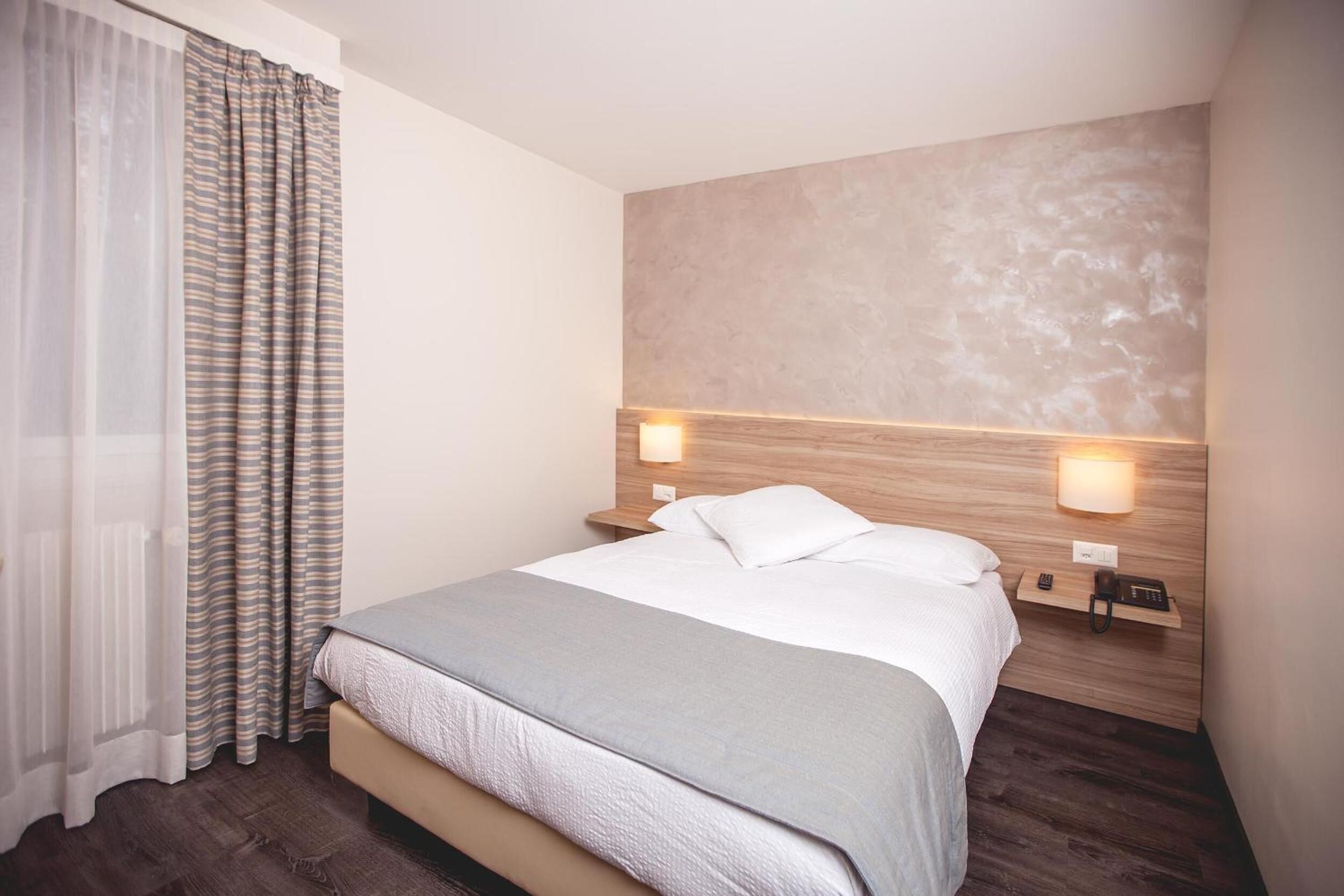 Una habitación de hotel moderna con una cama grande, dos mesitas de noche con lámparas, un cabecero de madera y una ventana con cortinas translúcidas.