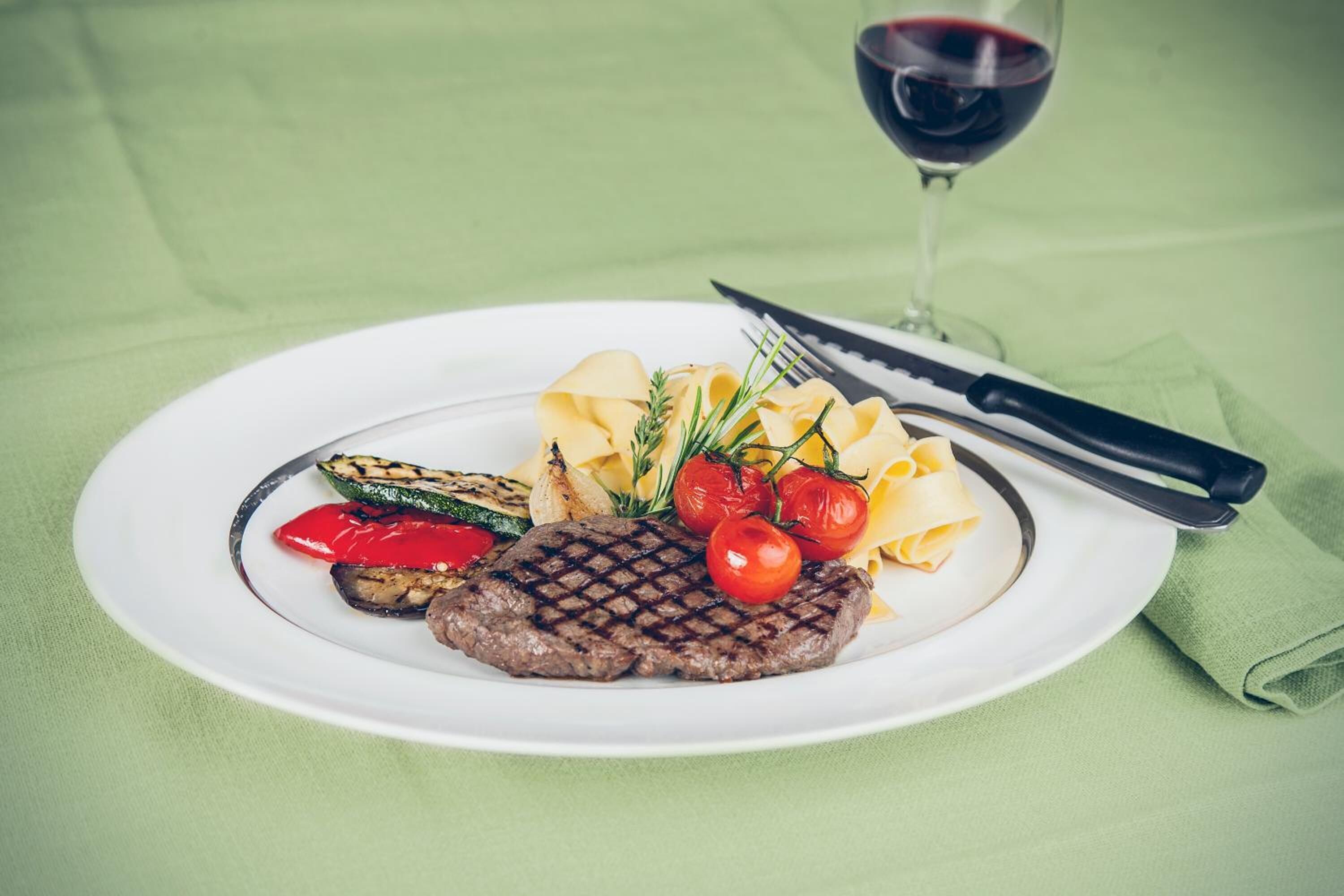 Een bord met een gegrilde steak, kersen tomaten, gegrilde groenten en pasta, vergezeld van een glas rode wijn.