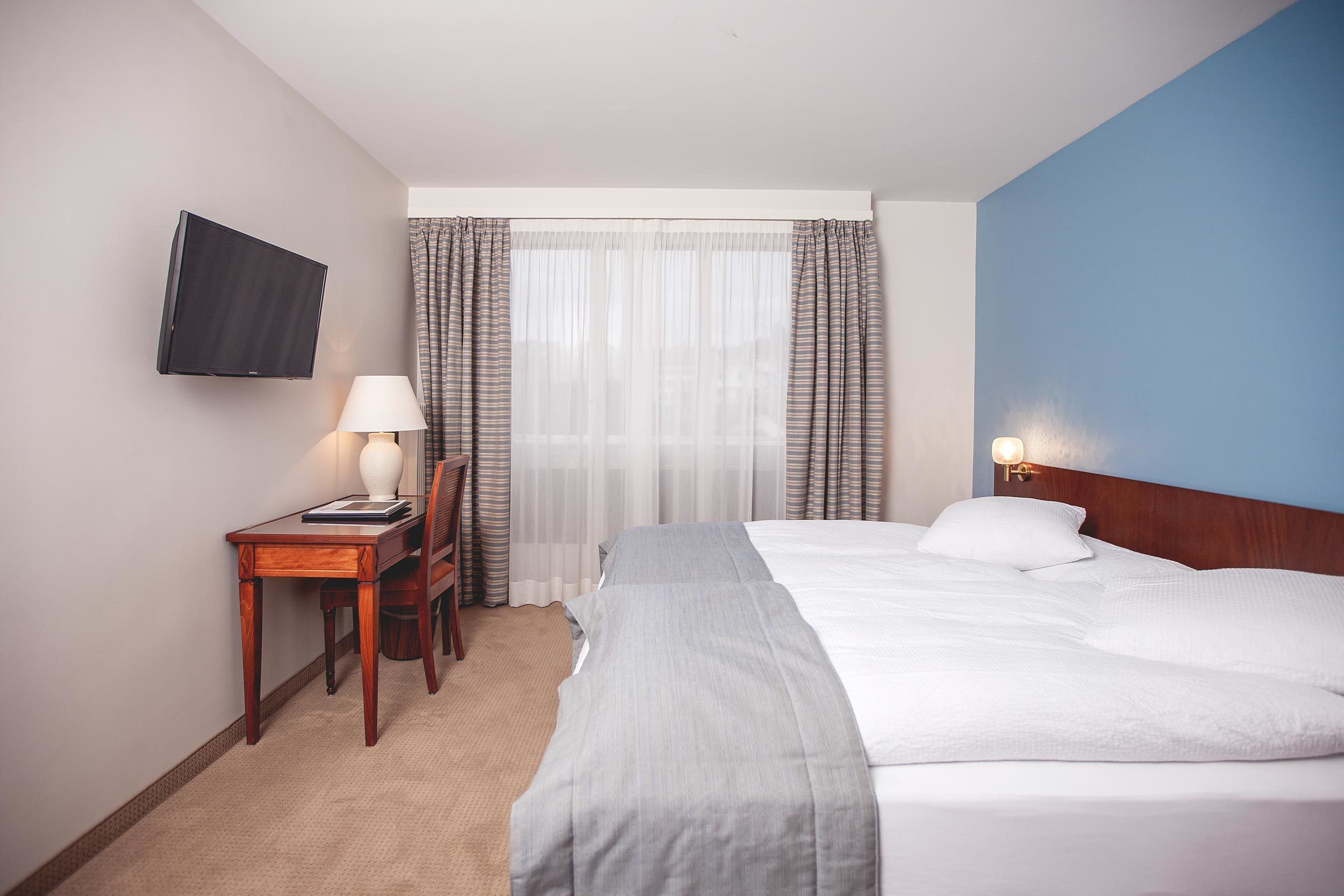 Una habitación de hotel con una cama, un escritorio con lámpara, una silla, un televisor montado en la pared y una ventana con cortinas.