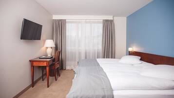 Una habitación de hotel con una cama, un escritorio con lámpara, una silla, un televisor montado en la pared y una ventana con cortinas.