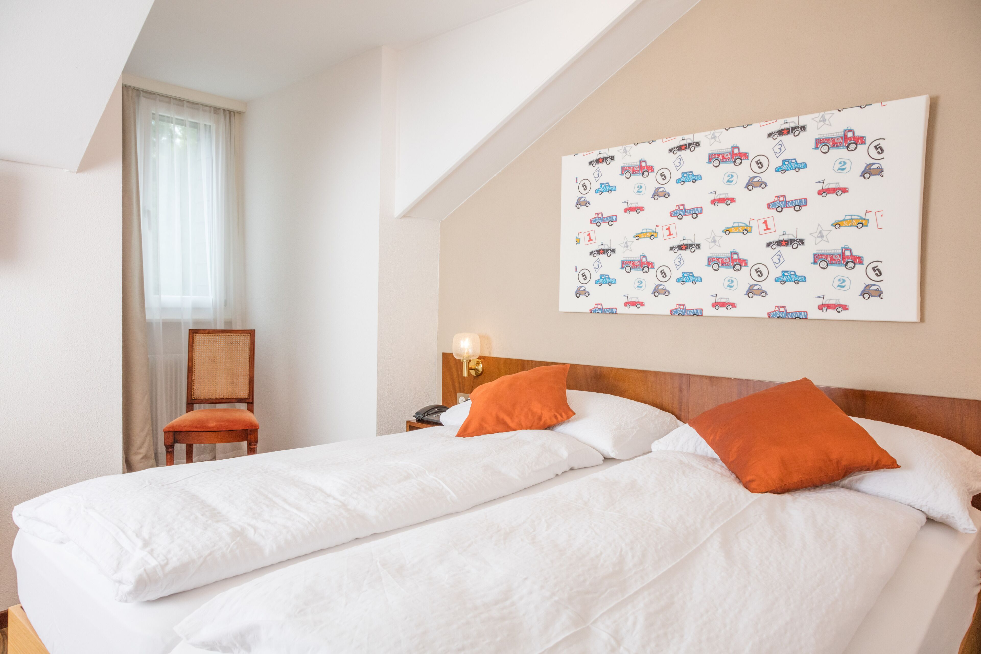 Quarto de hotel com uma cama grande, duas almofadas, uma mesa de cabeceira, uma luminária, uma cadeira e uma parede com padrão de carros infantis.