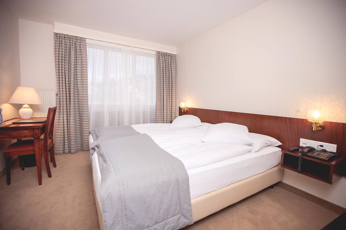 A hotel room with a bed, a desk with a lamp, a chair, a nightstand with a phone, and a window with curtains.