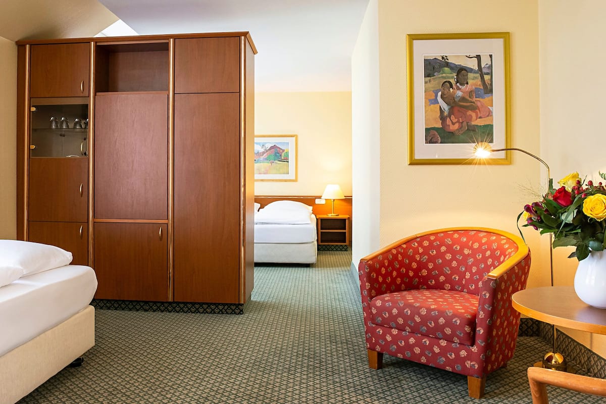 A hotel room with a bed, a wardrobe, a chair, a table with a vase, and a painting on the wall.