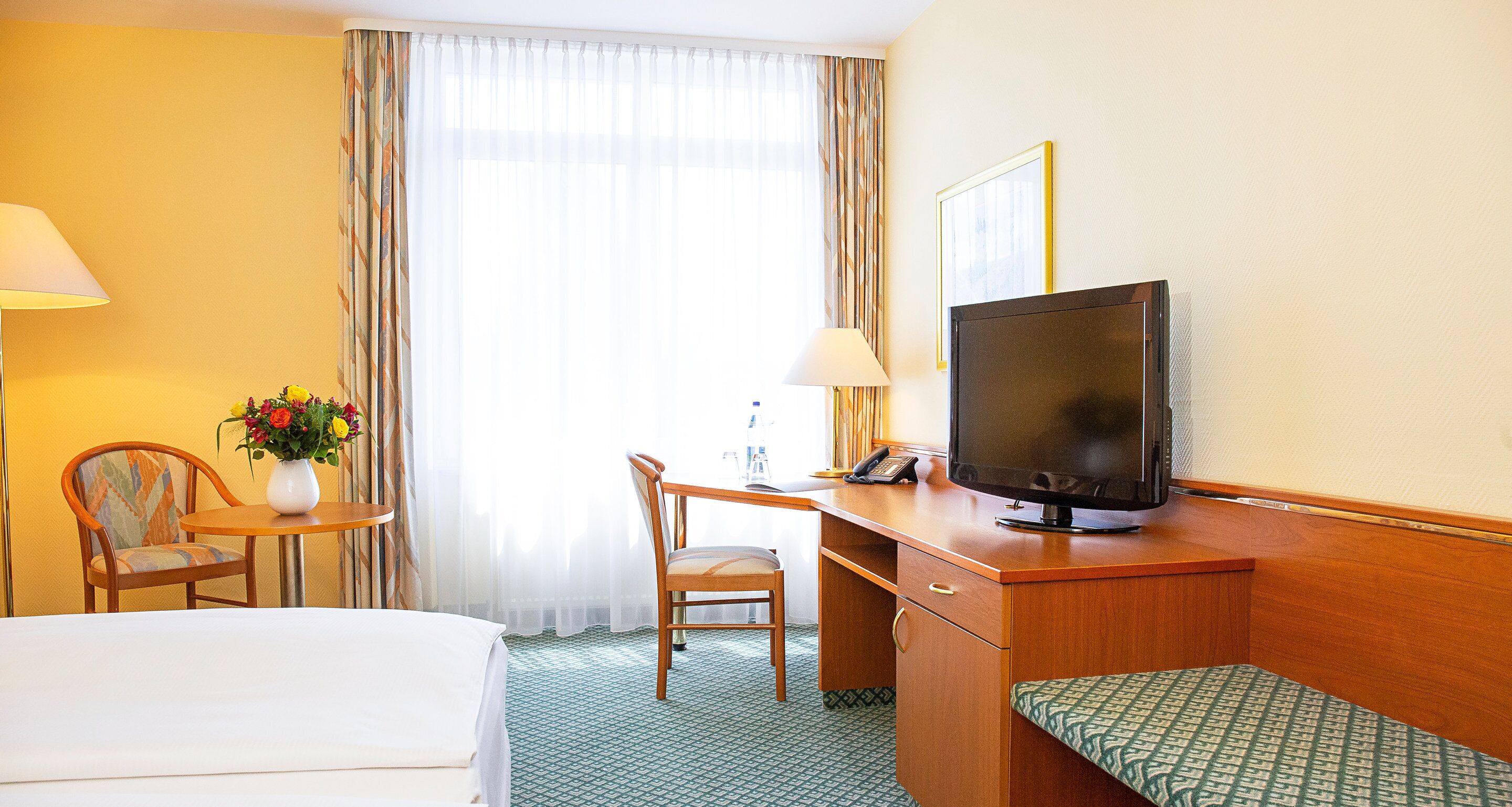 A hotel room with a bed, a desk with a computer, a chair, a TV, and a window with curtains.