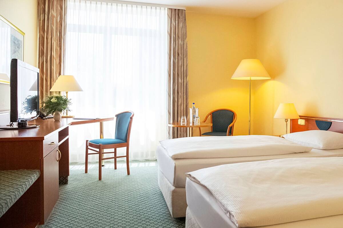 A hotel room with two beds, a desk with a computer, a chair, a lamp, and a painting on the wall.