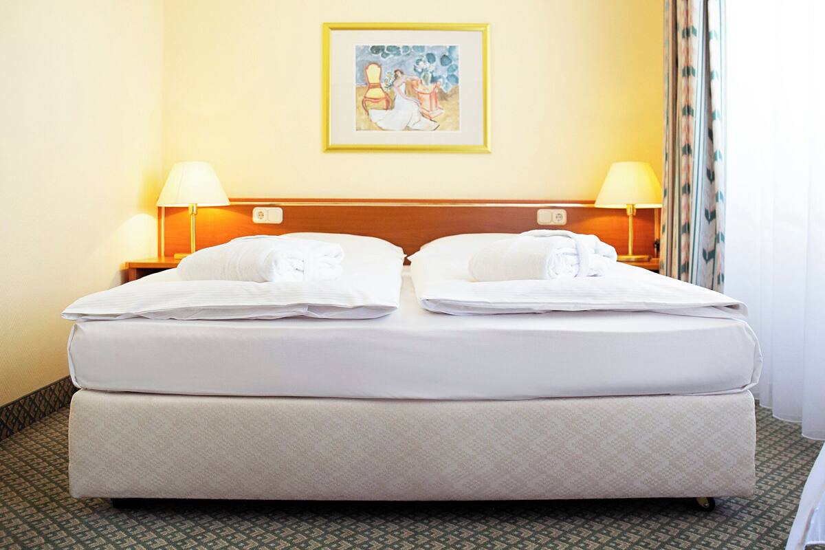 A hotel room with a double bed, two bedside lamps, a painting on the wall, and a patterned carpet.