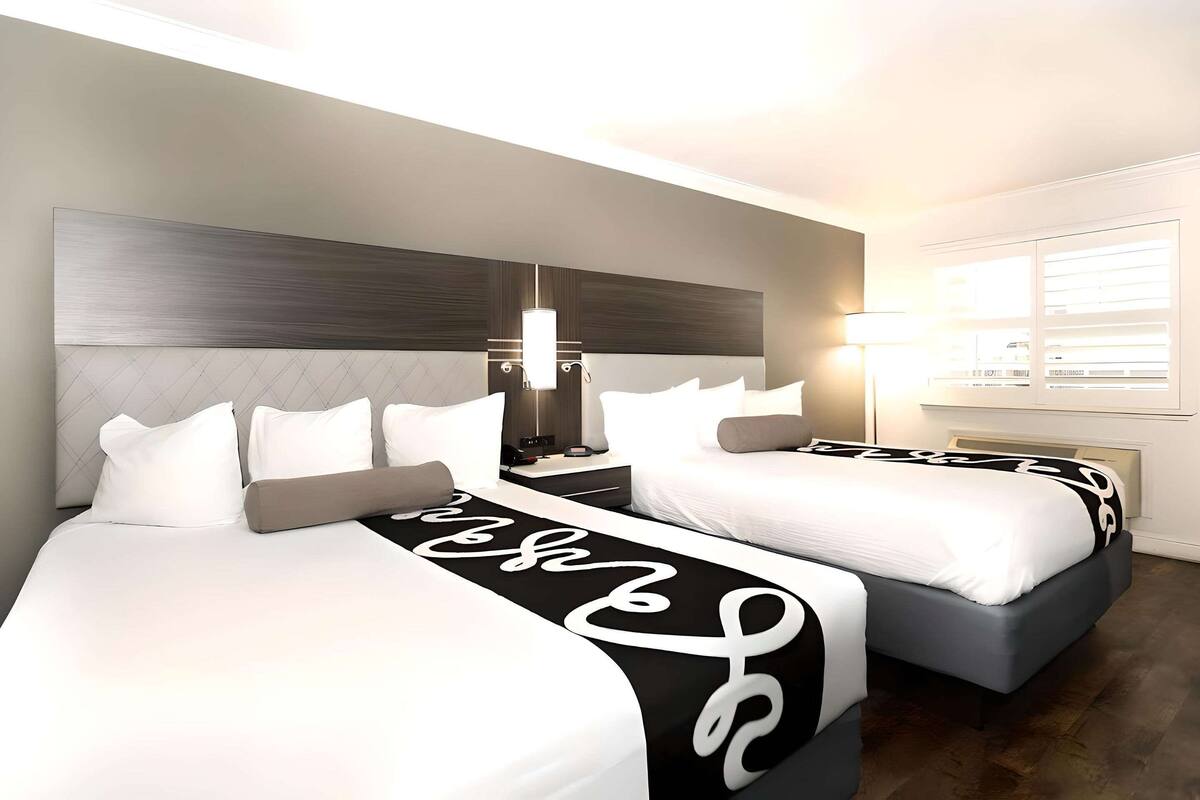 A hotel room with two beds, a modern headboard, bedside lamps, and a large window with blinds.