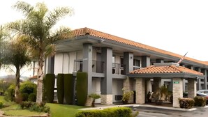 A multi-story hotel with a tiled roof, a covered entrance, and a well-maintained garden with palm trees and shrubs.