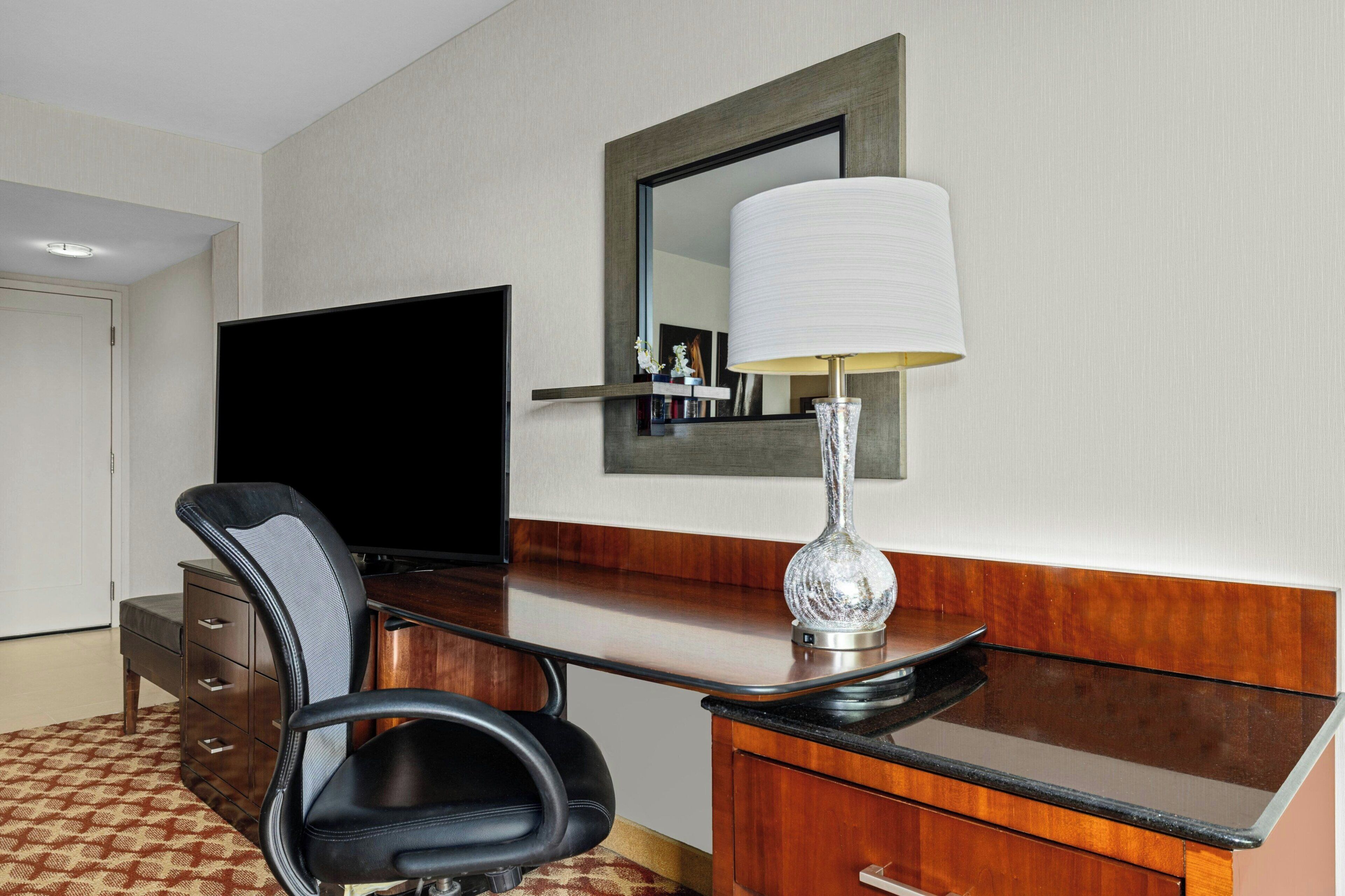 A hotel room with a desk, chair, television, and a lamp.