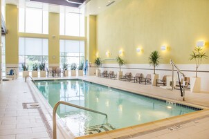 Una piscina interior con agua cristalina, iluminada por grandes ventanales, rodeada de sillones de descanso y plantas en macetas.