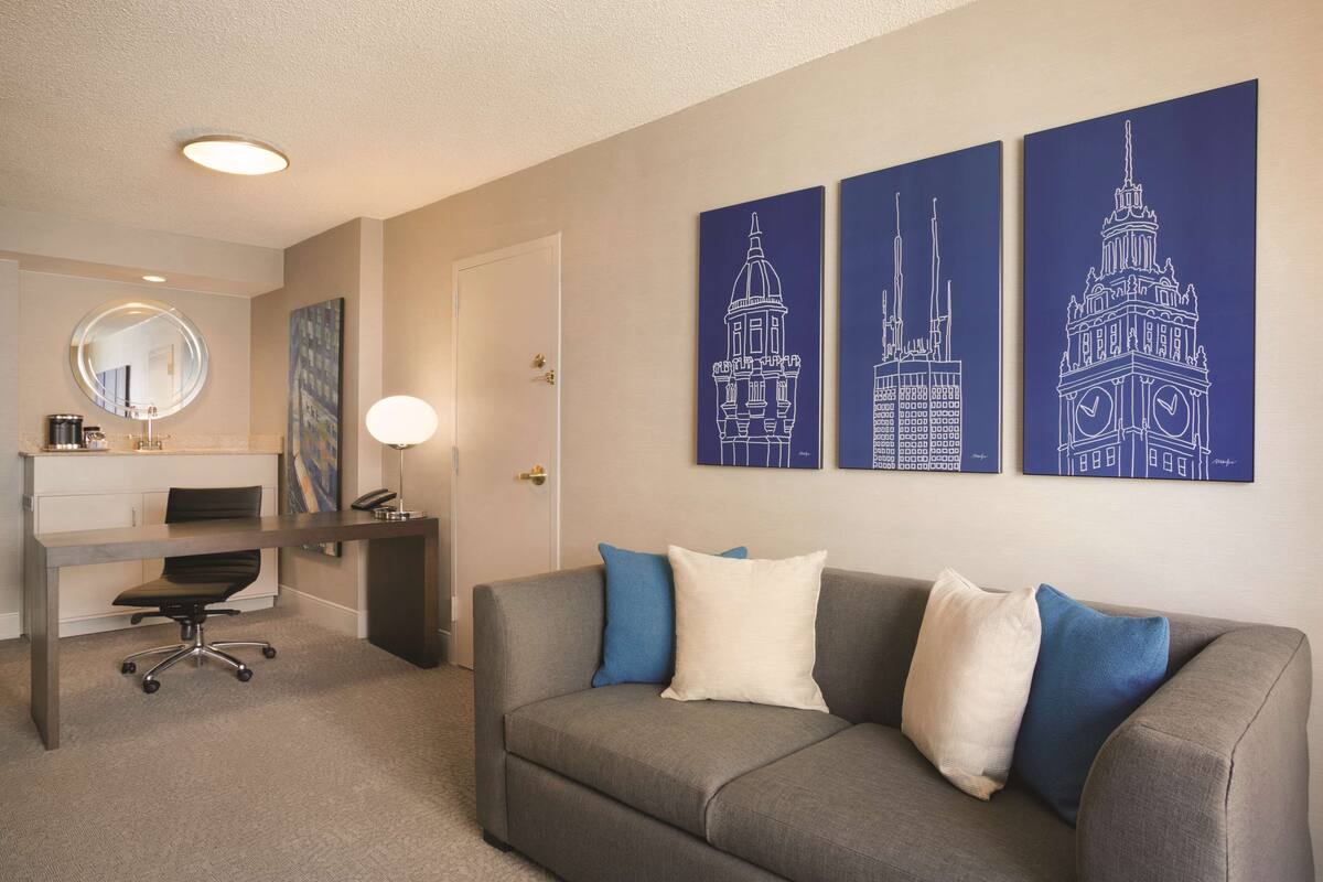 A modern hotel room with a grey sofa, a desk, and a chair. There are three wall art pieces depicting architectural structures.