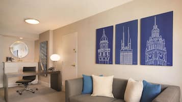 A modern hotel room with a grey sofa, a desk, and a chair. There are three wall art pieces depicting architectural structures.