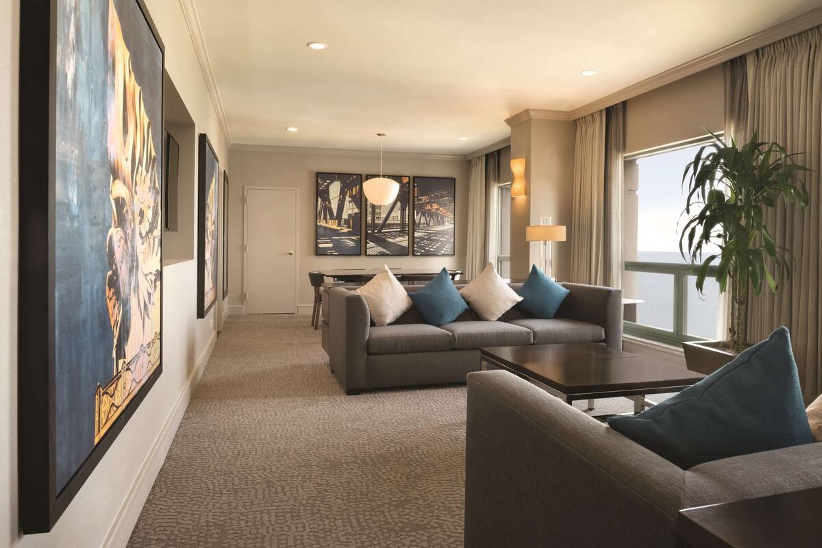 A modern hotel room with a grey sofa set, a coffee table, and a large window with curtains.