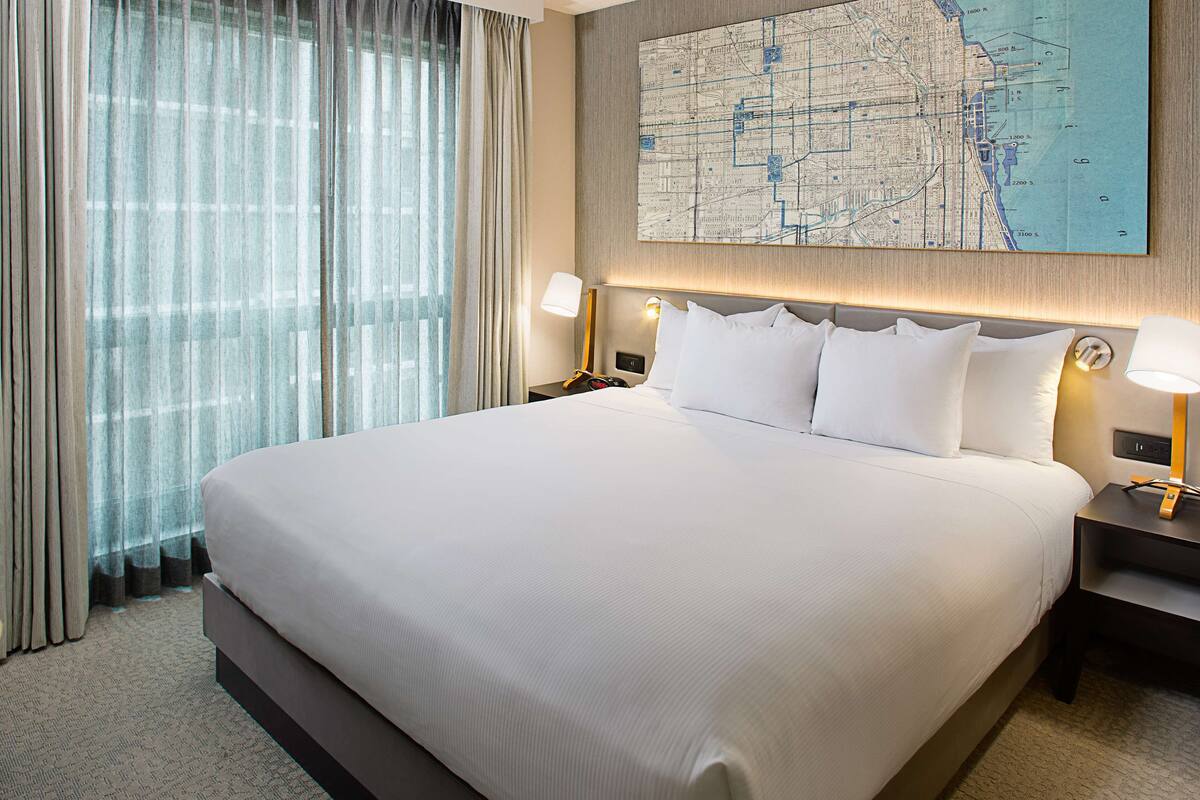 A large bed with white linens, two bedside tables with lamps, a large wall map, and sheer curtains.