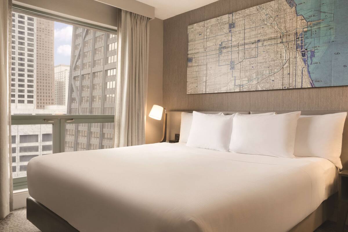 A hotel room with a large bed, a map on the wall, and a view of buildings through the window.