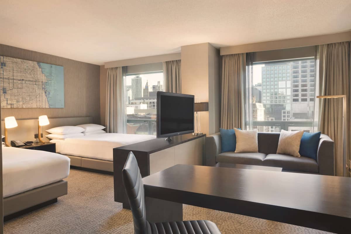 A hotel room with a bed, a desk with a TV, a sofa, and a city view through large windows.