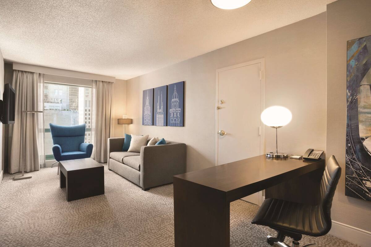 A hotel room with a large window, a sofa, a desk with a chair, and artwork on the wall.
