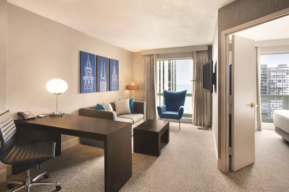 A hotel room with a desk, chair, sofa, and armchair. There are two artworks on the wall, a TV mounted on the wall, and a window with curtains.
