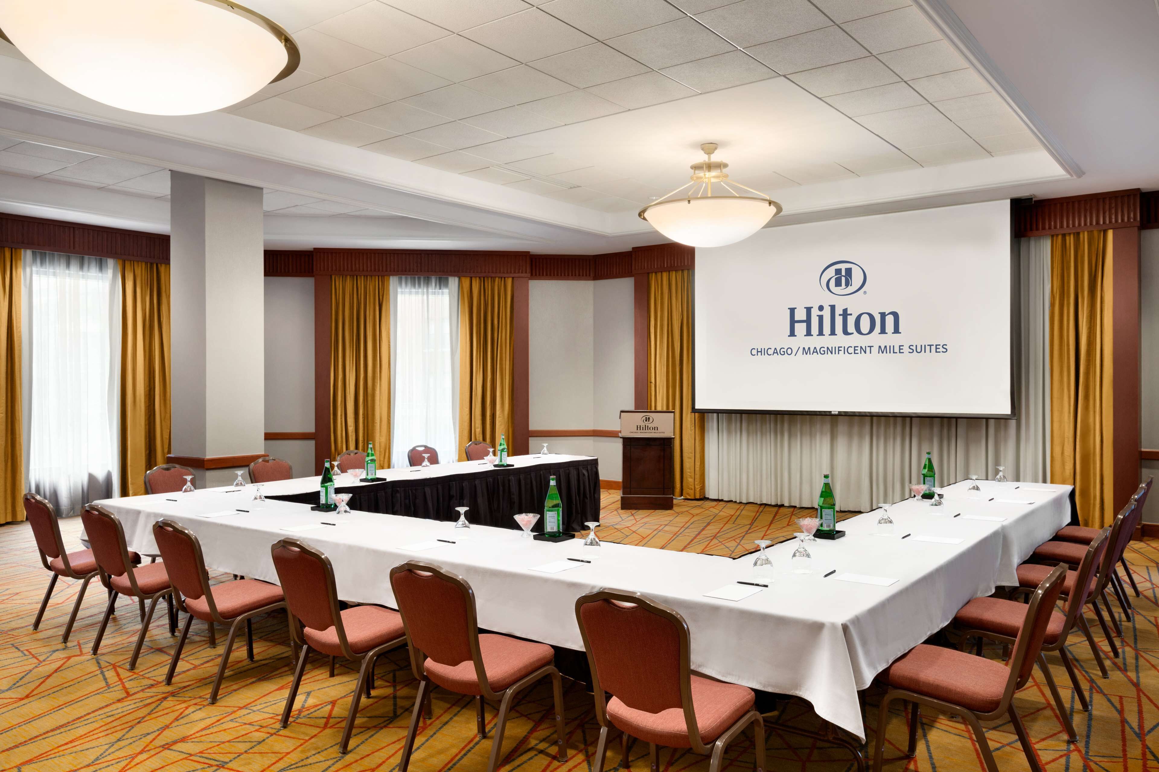 Un salón de conferencias con una larga mesa, sillas, una pantalla de proyector y un atril con el logotipo de Hilton.
