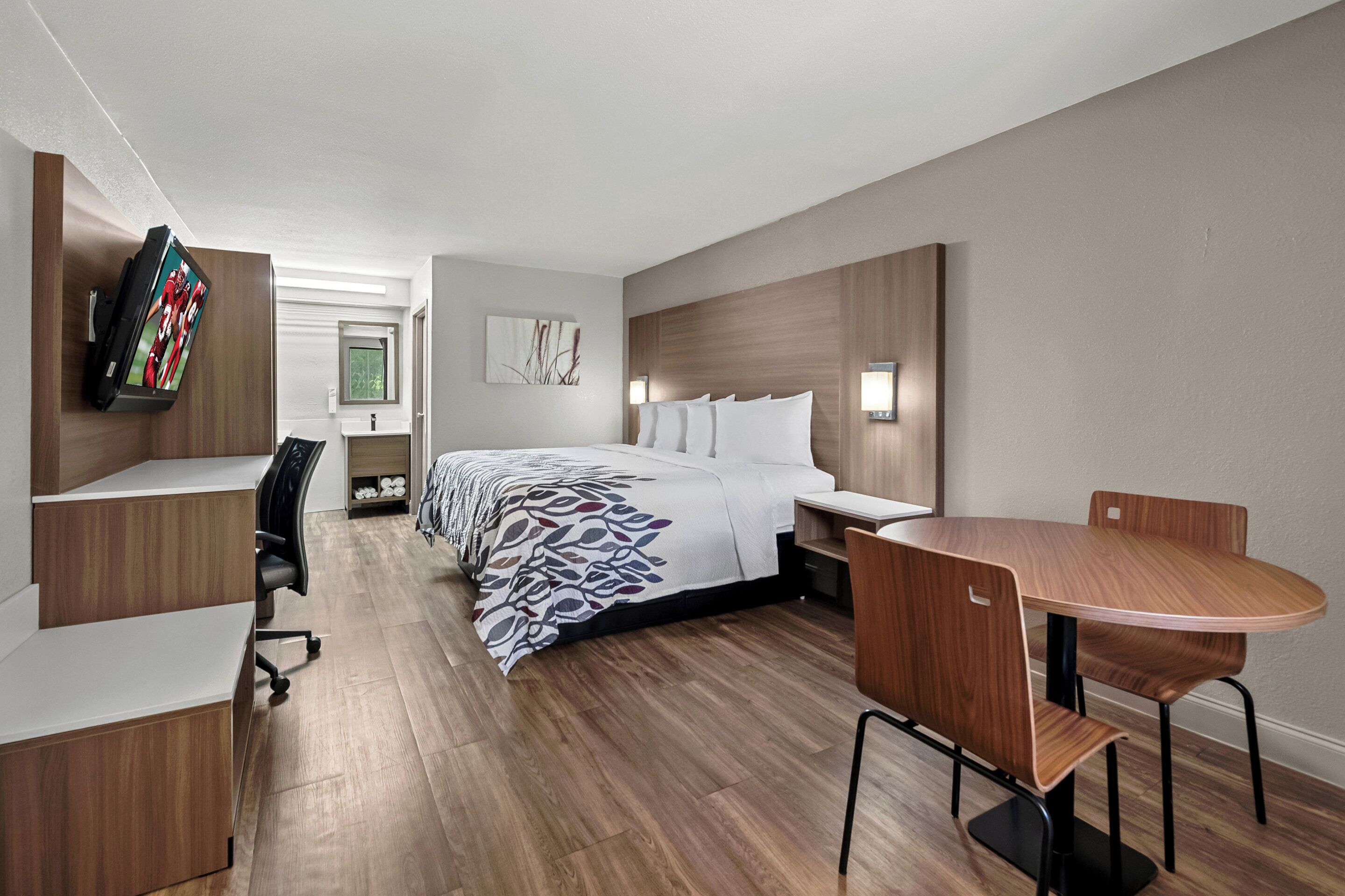 A modern hotel room with a neatly arranged bed, a desk with a chair, a dining table with chairs, and a flat-screen TV mounted on the wall.