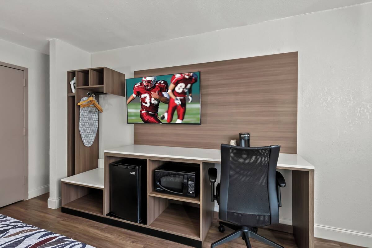 A compact room with a built-in desk, a microwave, and a TV displaying a sports game.