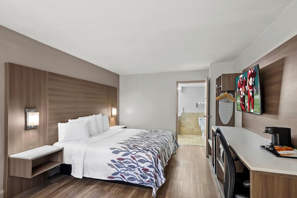 A hotel room with a bed, bedside table, a desk with a chair, a TV, and a shower area. - Red Roof Inn Sanford (Sanford)