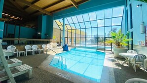 Una piscina interior con paredes de vidrio transparente, rodeada de sillas y mesas blancas, y un techo de madera.