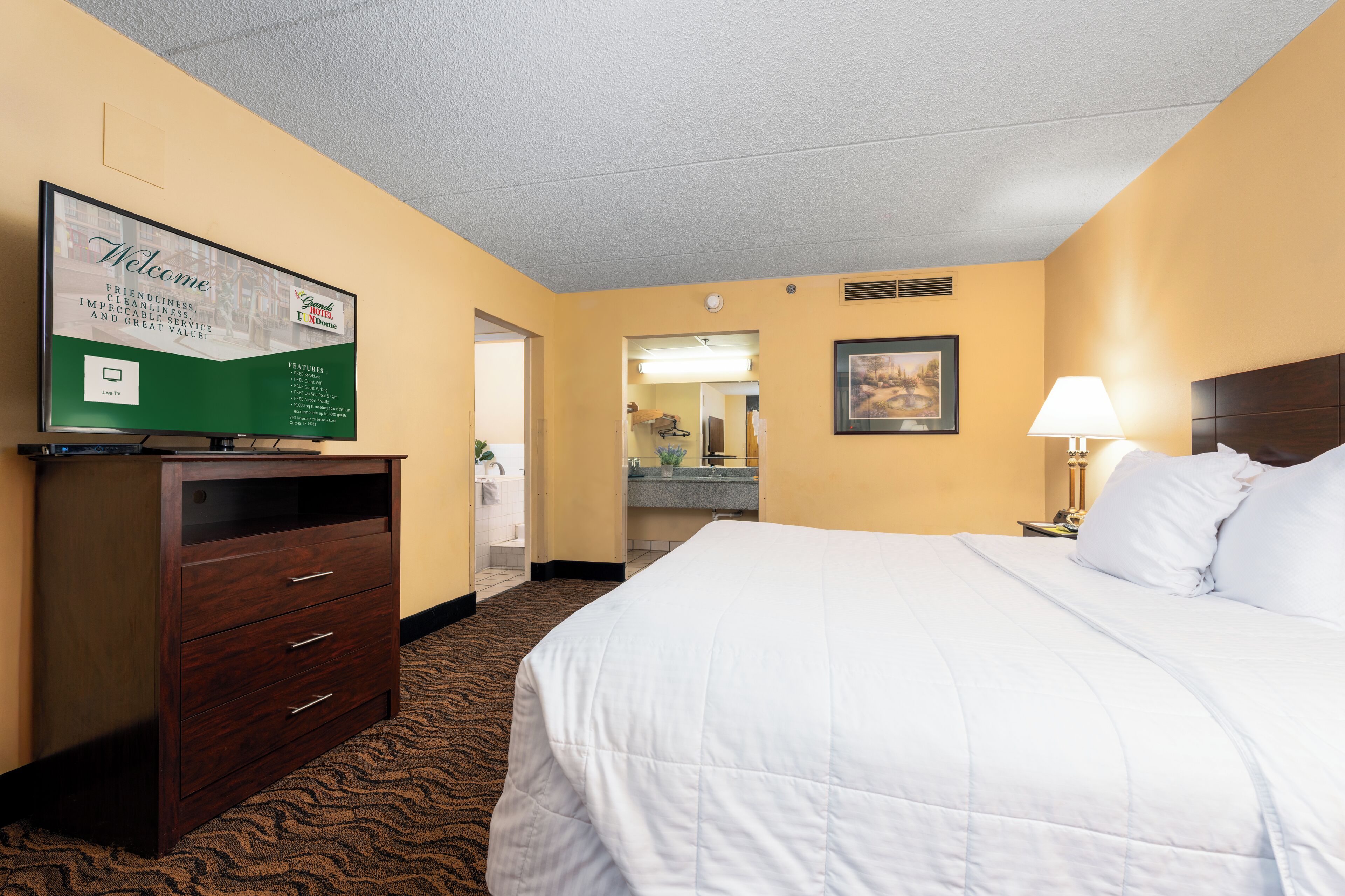 A hotel room with a bed, a dresser, a television displaying a welcome message, a bathroom visible through an open door, and a nightstand with a lamp.