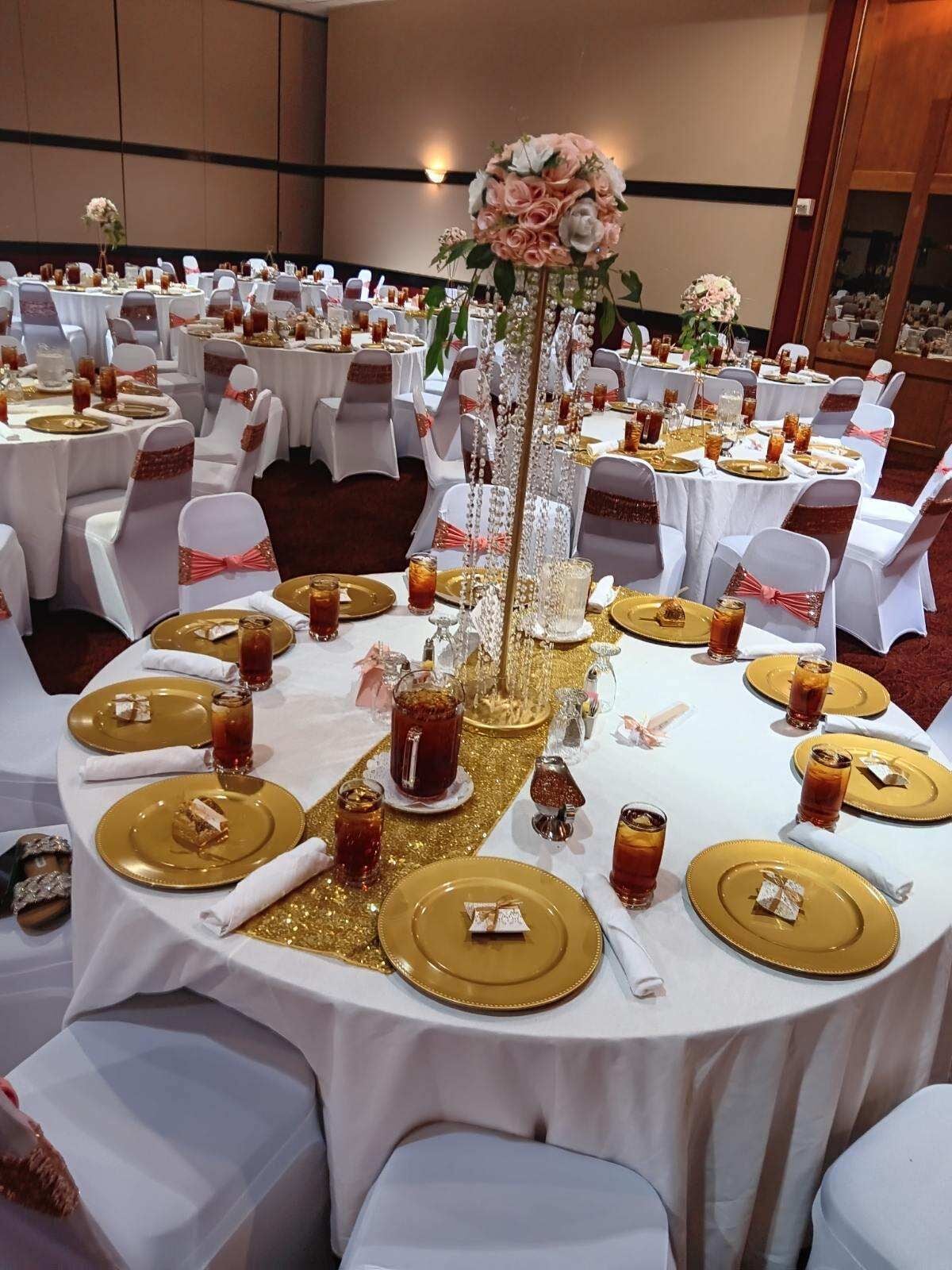 A banquet hall with round tables set for a formal event, featuring gold and white tablecloths, white chairs with pink sashes, and floral centerpieces.