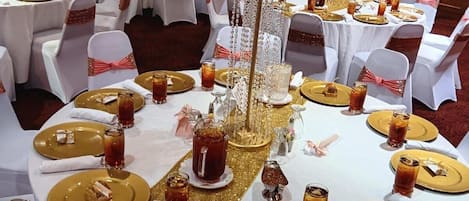 A banquet hall with round tables set for a formal event, featuring gold and white tablecloths, white chairs with pink sashes, and floral centerpieces.