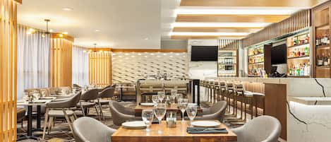 A modern restaurant with a long dining area, bar, and a TV screen.