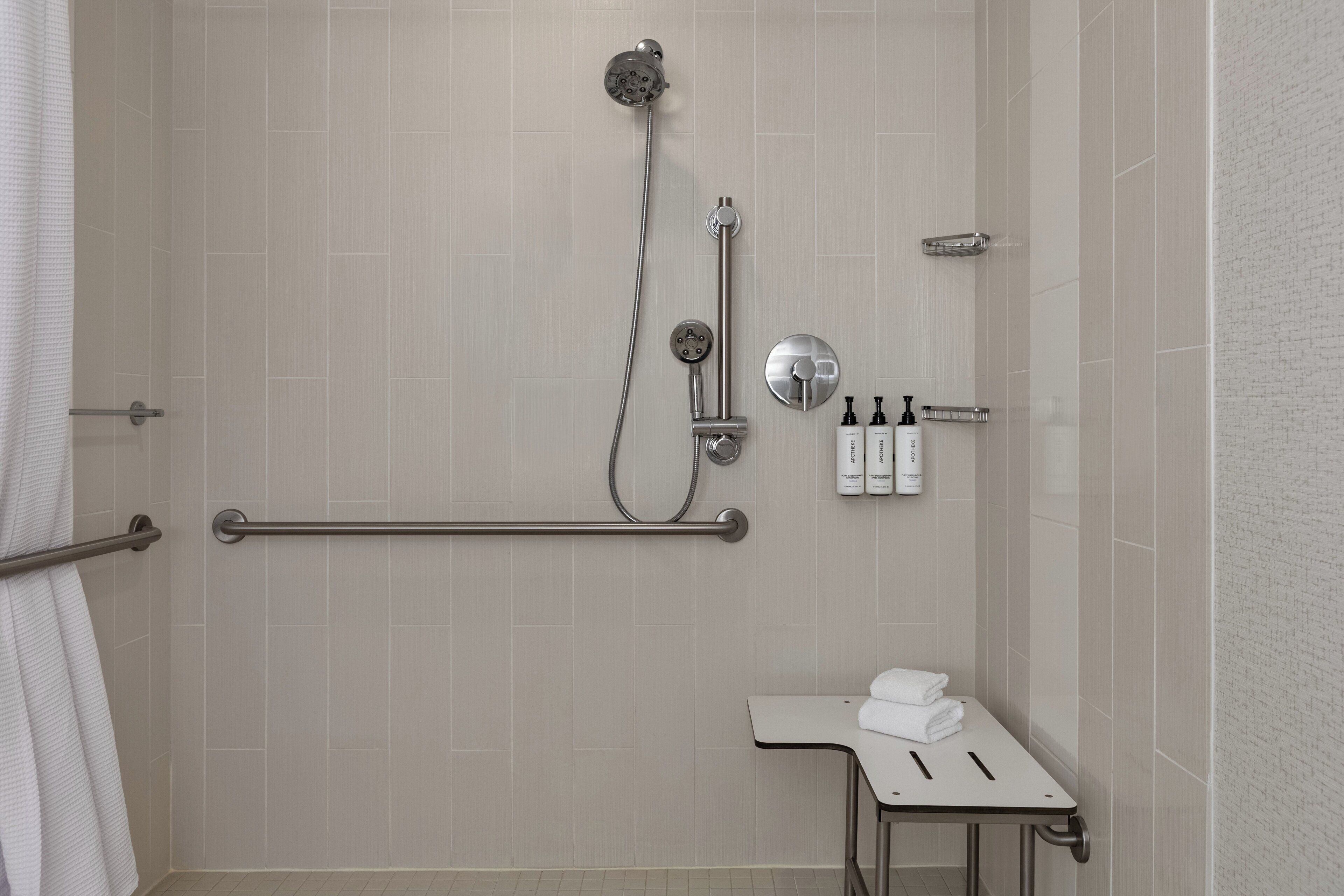 Un espace douche équipé d’un pommeau de douche à main, d’un robinet mural et d’une barre d’appui. On y trouve deux bouteilles de shampoing et une serviette blanche posées sur une petite table.