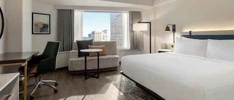 Habitación de hotel con una cama grande, un escritorio, una silla, una mesita y vistas a la ciudad a través del ventanal.