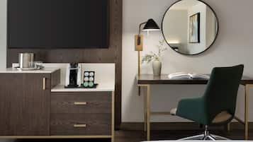 A hotel room with a flat-screen TV, a coffee machine, a desk with a chair, a vase with a plant, and a round mirror on the wall.