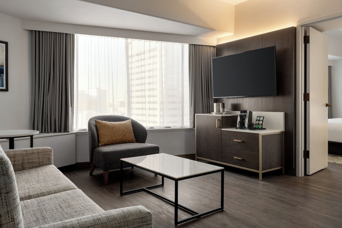 A modern hotel room with a sofa, a coffee table, a TV, and a desk with drawers.