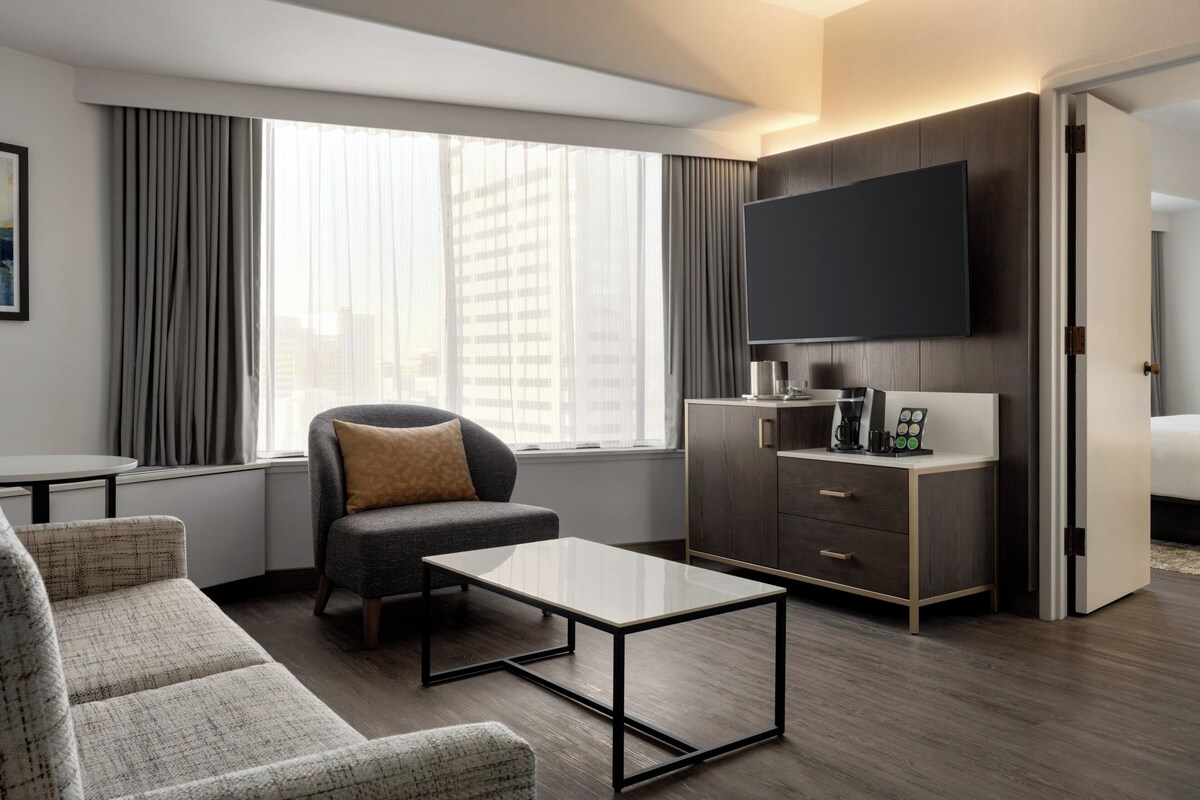 A modern hotel room with a sofa, a coffee table, a TV, and a desk with drawers.