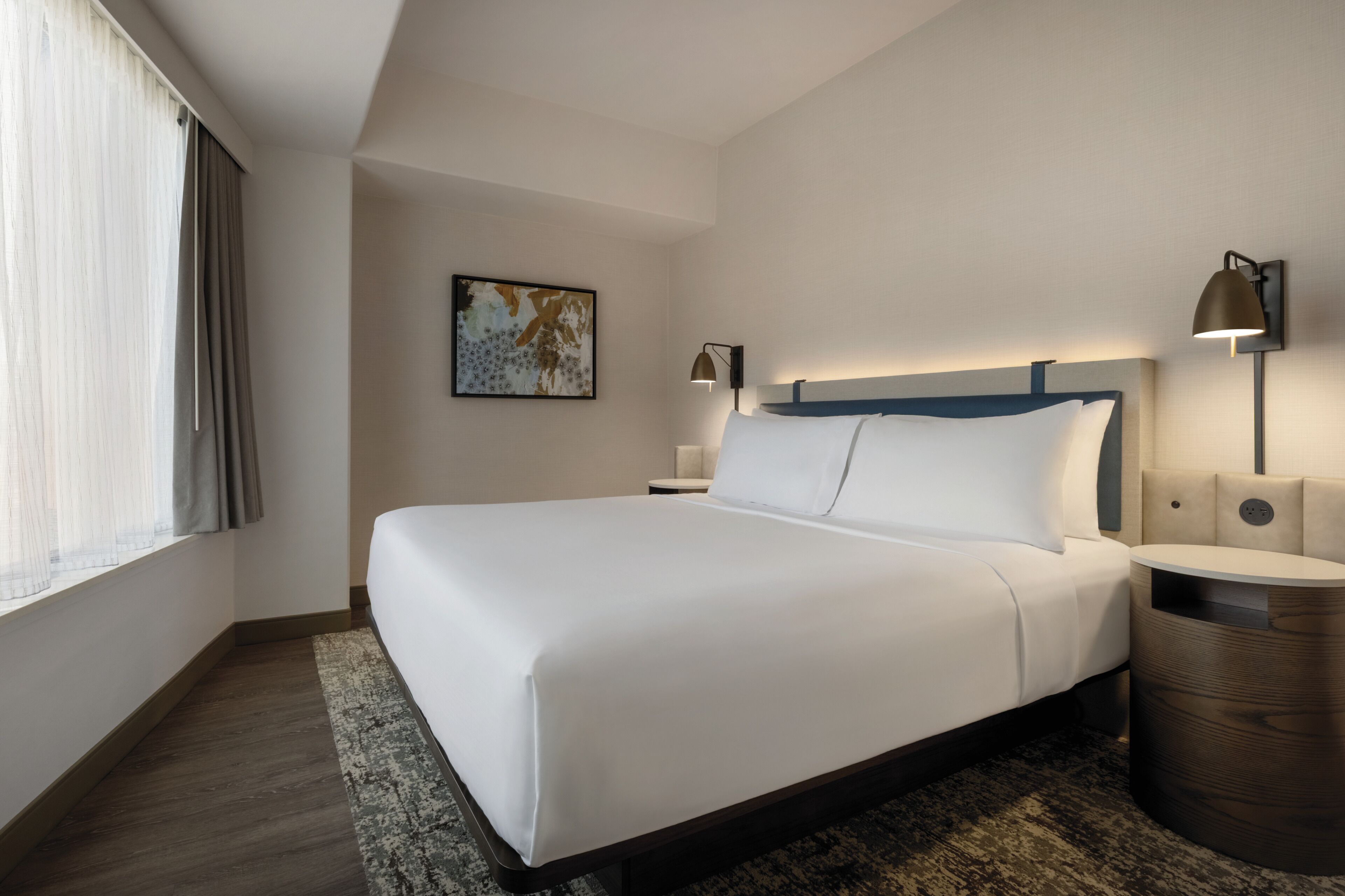 Una habitación de hotel moderna con una cama grande, una mesita de noche, una lámpara de pared y una ventana con cortinas.