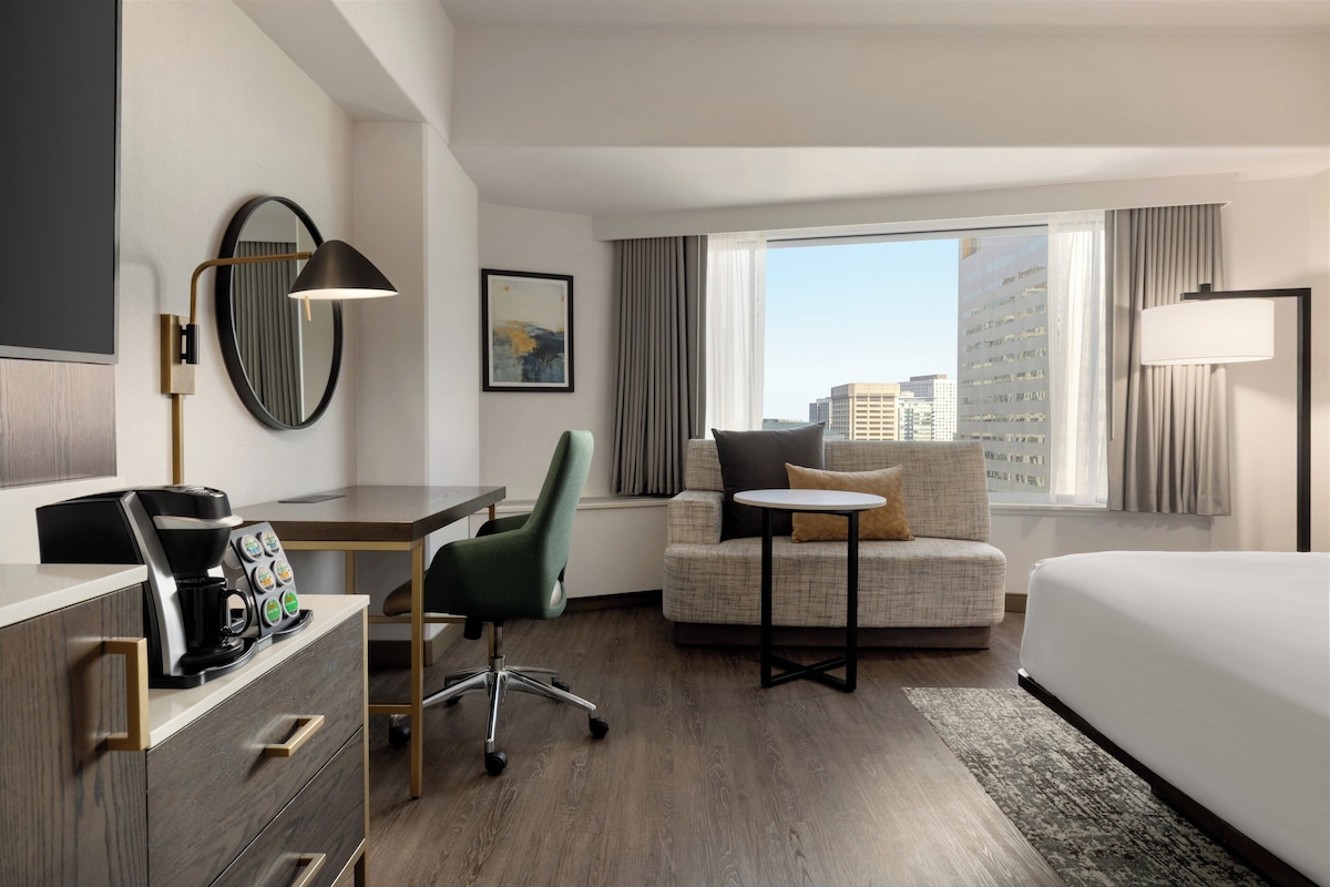 A hotel room with a bed, a desk with a coffee maker, a chair, a sofa, a large window with a city view, and a wall-mounted lamp.